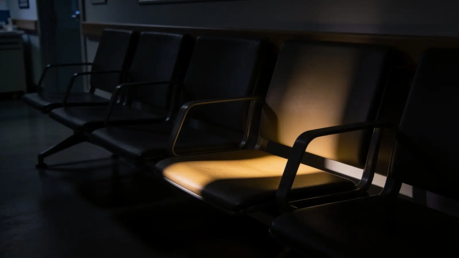 Fila de sillas vacías en la sala de espera de una clínica, con un haz de luz cálida sobre un solo asiento.