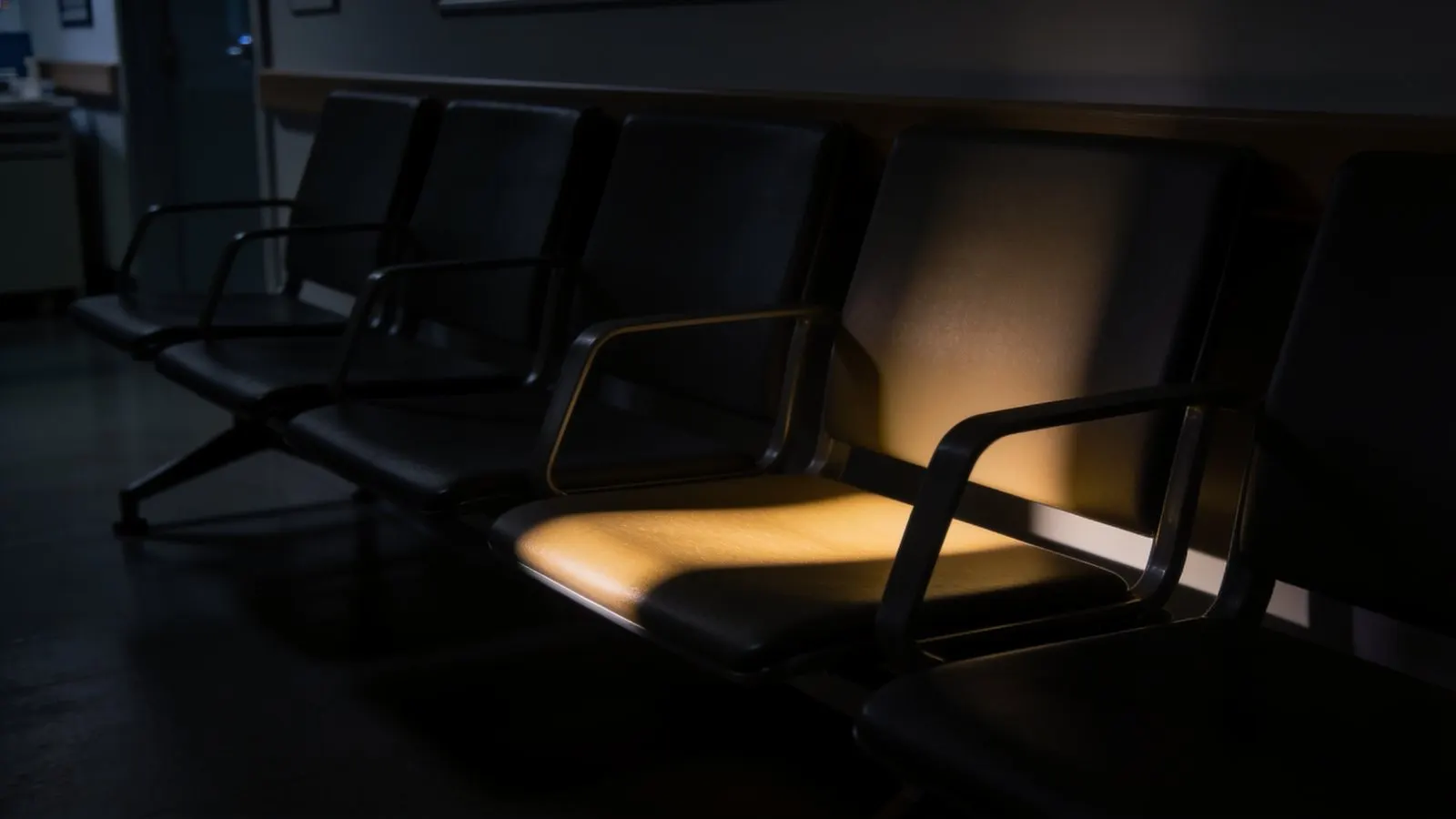 Fila de sillas vacías en la sala de espera de una clínica, con un haz de luz cálida sobre un solo asiento.